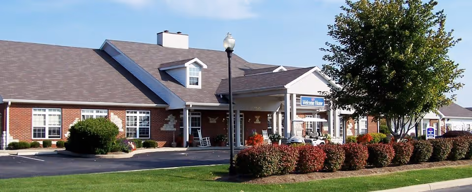 Exterior view of a single-story brick building with white trim, a covered entrance with a 'Welcome Home' sign, surrounded by neatly trimmed bushes, a tree, and a well-maintained lawn under a clear blue sky.