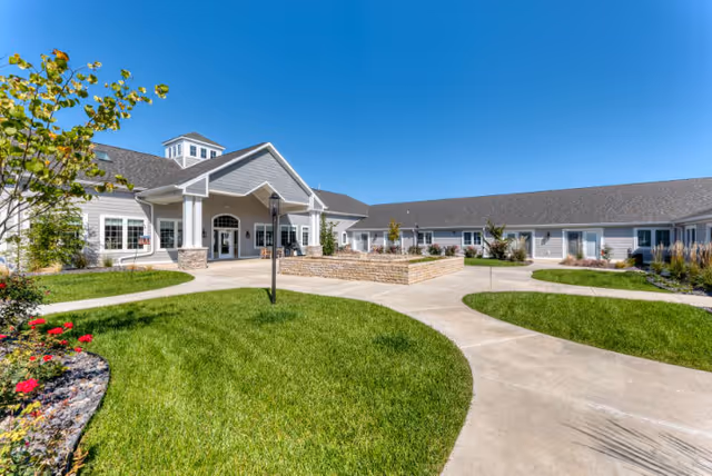 Exterior view of a single-story senior living facility building with a covered entrance, surrounded by well-maintained green lawns, flower beds, and paved walkways under a clear blue sky.