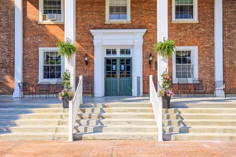 Front entrance of a brick building with green double doors framed by white columns and decorative molding. There are concrete steps leading up to the entrance with white railings on both sides, potted plants with flowers and hanging ferns on either side of the door, and black metal chairs and tables placed near the windows.