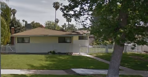 A single-story residence with a green lawn, white fence, and several trees.