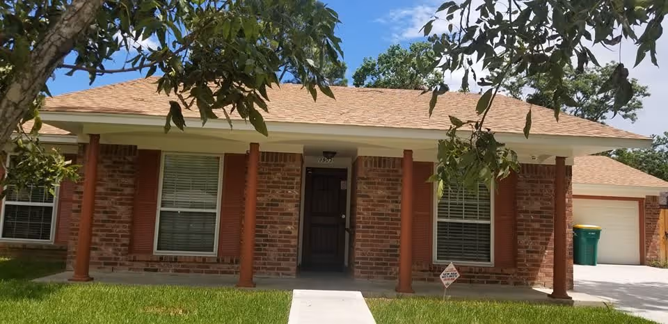 Front exterior view of a single-story brick house with a covered porch supported by four columns, two windows with closed blinds, a front door, a driveway leading to a garage, and a green trash bin near the garage. There is a tree branch partially visible at the top of the image and a small lawn in front of the house.