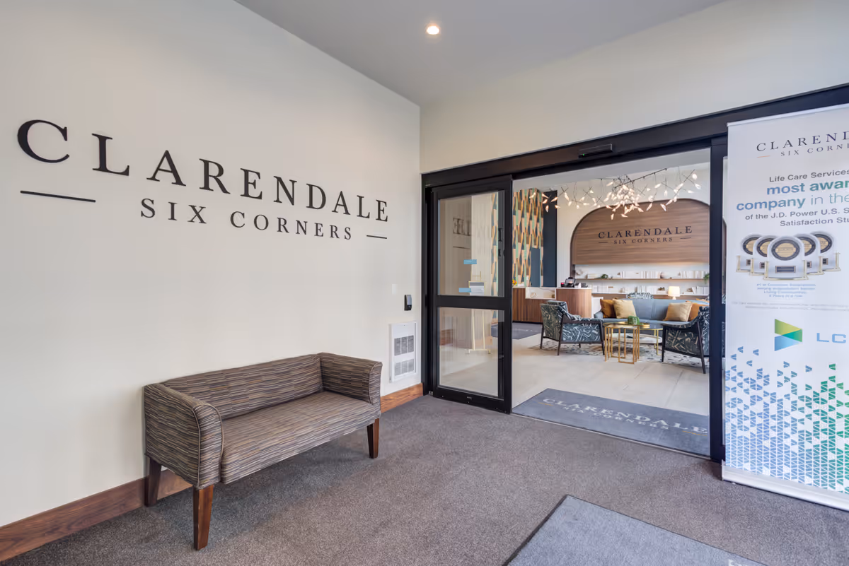 Entrance area of Clarendale Six Corners facility showing a small striped bench against a wall with the facility name on it. Through glass doors, a reception area with chairs, a table, and a decorative wall with the Clarendale Six Corners logo is visible. A standing banner with information about the facility is on the right side.