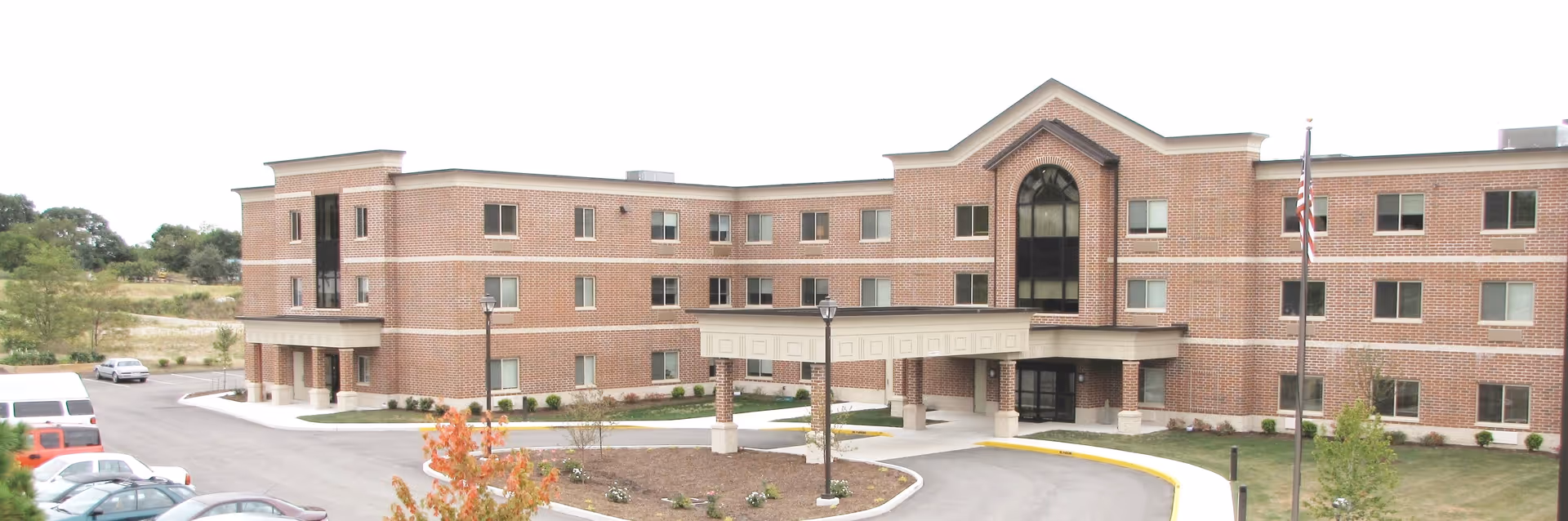 Front exterior view of a three-story brick senior living facility with multiple windows, a covered entrance, a flagpole with an American flag, and a parking area with several cars.