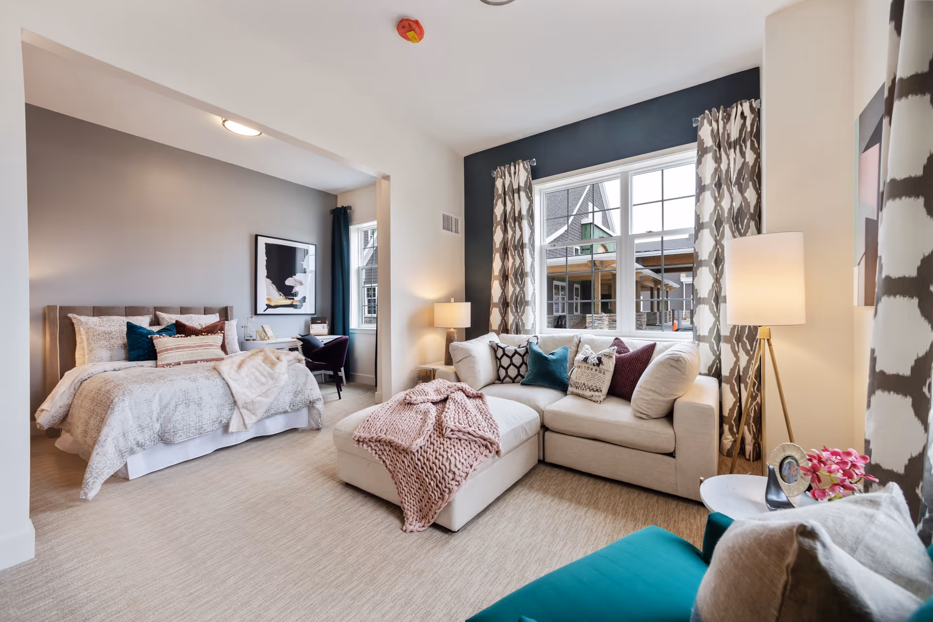 A bright and cozy living space featuring a beige sectional sofa with multiple decorative pillows and a pink knitted throw blanket. The room has large windows with patterned curtains allowing natural light to enter. Adjacent to the living area is a bedroom with a neatly made bed, decorative pillows, a desk, and a chair. The walls are painted in neutral tones, and the floor is carpeted in a light beige color. A floor lamp and a small side table with a photo frame and flowers complete the scene.