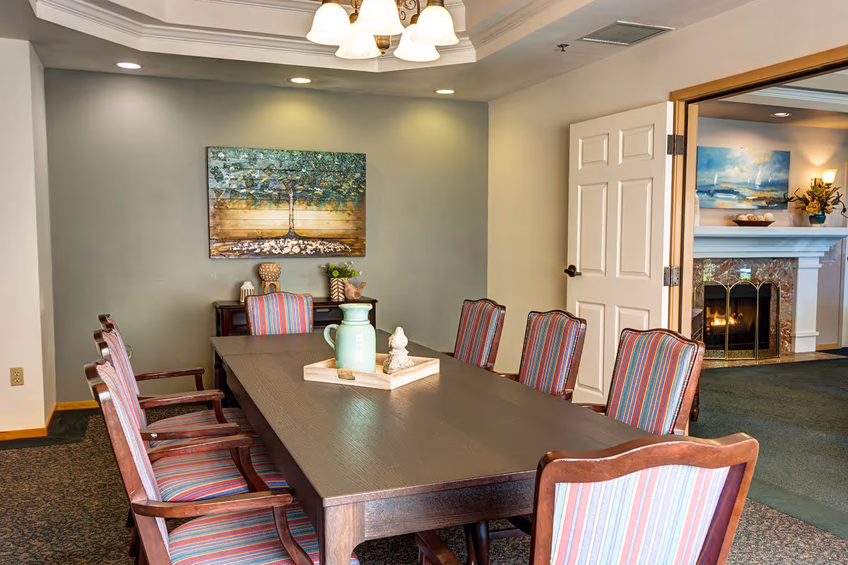 A dining room with a long wooden table surrounded by eight striped upholstered chairs. On the table is a wooden tray holding a green pitcher and a decorative item. The walls are painted light gray, with a painting of a tree hanging on one wall. A doorway leads to another room with a fireplace and a painting above it.