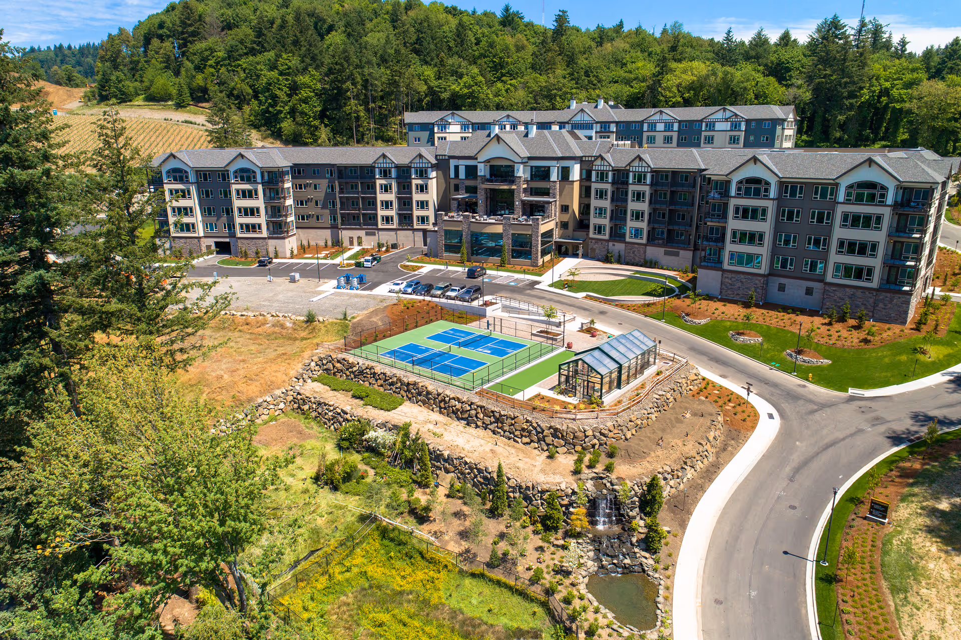 Aerial view of a large senior living facility named Touchmark in the West Hills surrounded by trees and greenery. The building is multi-story with numerous windows and balconies. In front of the building, there is a tennis or pickleball court and a small greenhouse structure. The facility is bordered by a curved road and landscaped areas with grass, shrubs, and a small waterfall feature.
