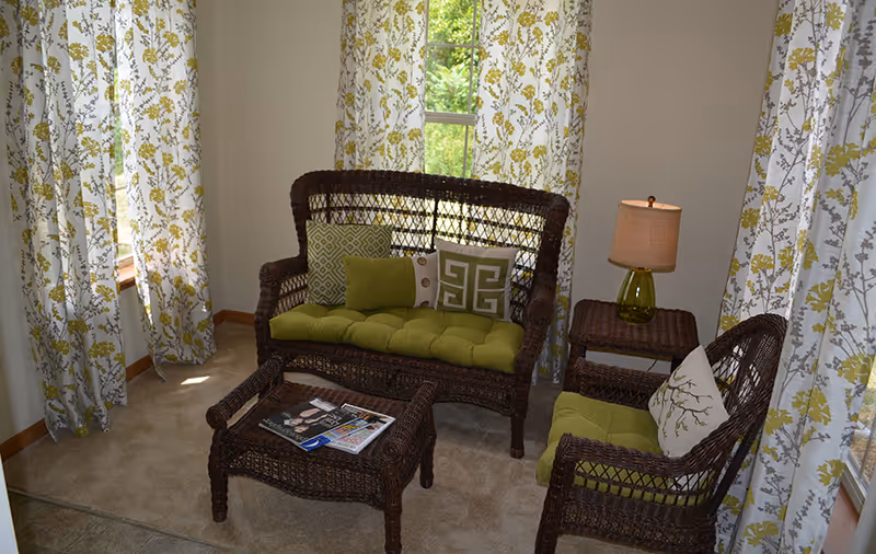 A cozy sitting area with dark wicker furniture including a loveseat, chair, coffee table, and side table. The loveseat and chair have green cushions and decorative pillows. A lamp with a green glass base and beige shade sits on the side table. The room has beige carpet and walls, with windows covered by white curtains featuring a green and yellow floral pattern.