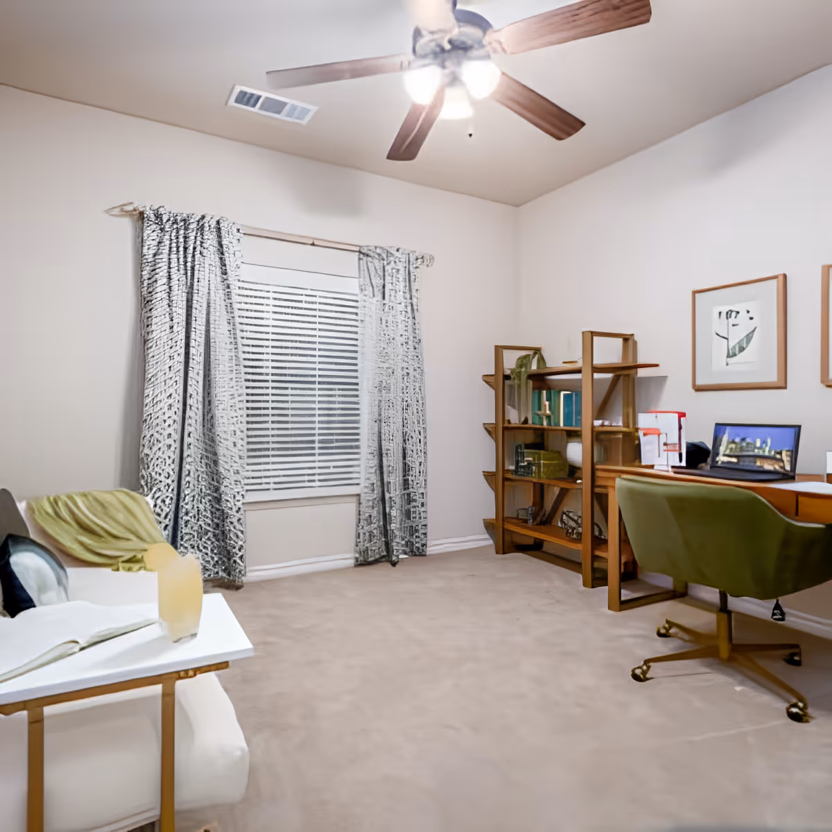 A cozy interior room with beige carpet and white walls. There is a ceiling fan with lights, a window with patterned curtains, a wooden bookshelf filled with books and decorative items, a wooden desk with a green swivel chair, and a white armchair with a green throw blanket and an open book on a side table.