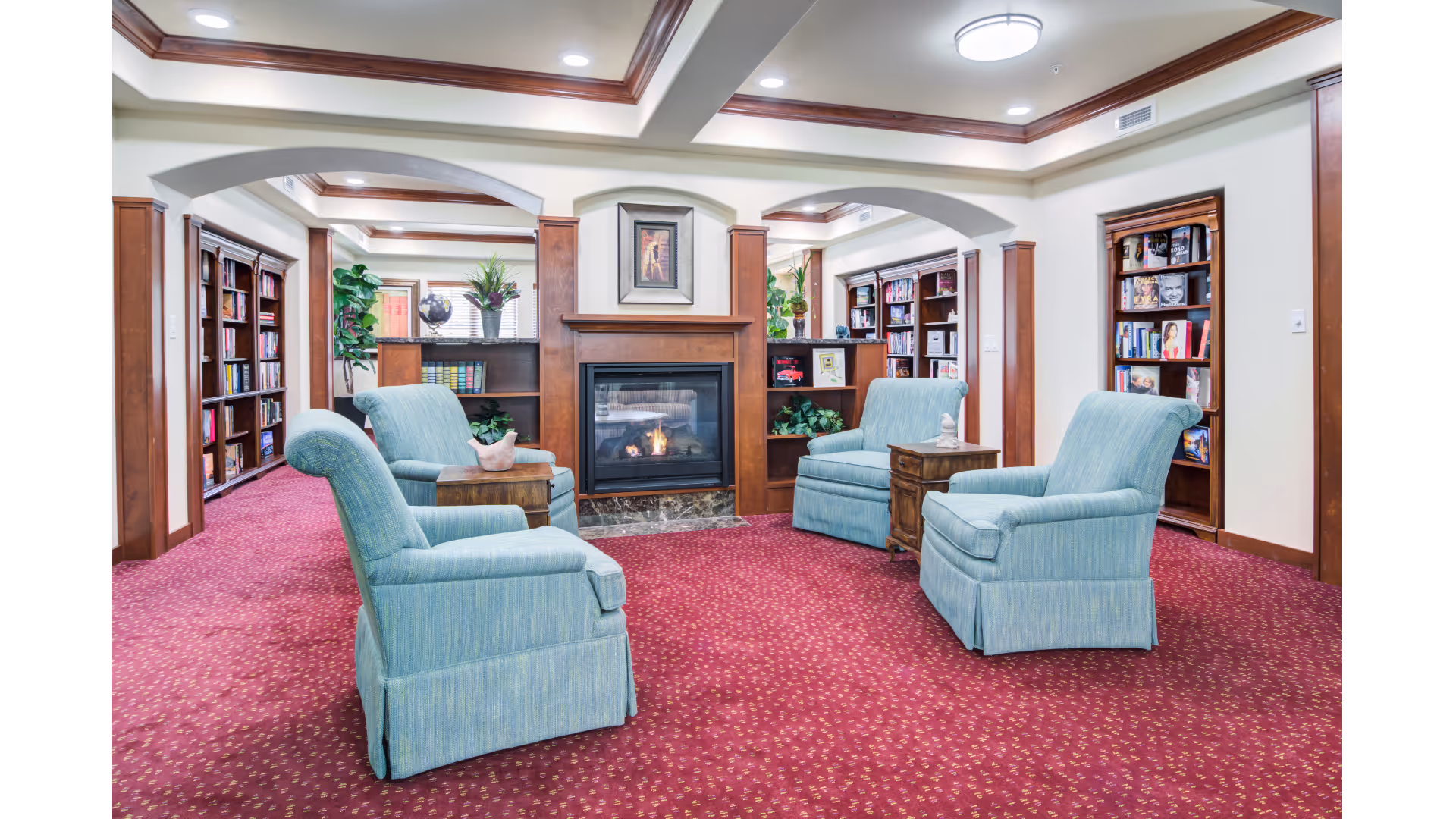 A cozy living room area with four light blue upholstered armchairs arranged around a wooden fireplace with a fire burning. The room has red patterned carpet, wooden trim, built-in bookshelves filled with books, and decorative plants. The ceiling features recessed lighting and wooden crown molding.
