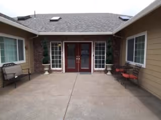 Outdoor courtyard area with a concrete floor, two sets of double glass doors with red frames, beige siding walls, two potted topiary plants flanking the doors, a bench on the left side, and two chairs with a small table on the right side.
