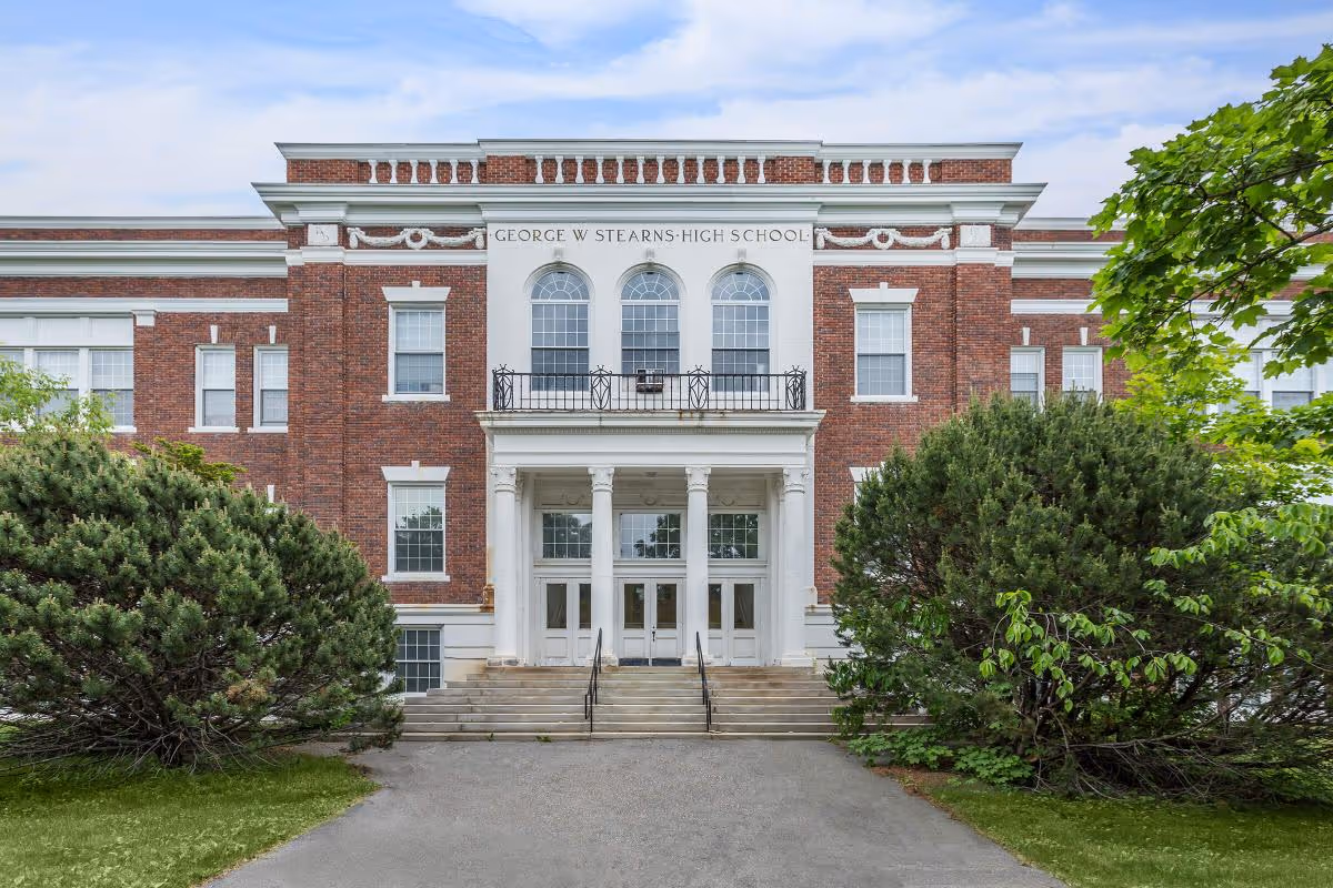 Red-brick neoclassical building front with columns, steps, and the inscription "GEORGE W STEARNS HIGH SCHOOL" above the main entrance, flanked by large shrubs.