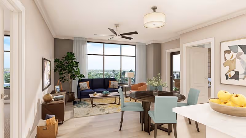 Bright open-plan living and dining area with a navy sofa, round dining table, large window with a scenic view, and a kitchen counter with a bowl of apples.