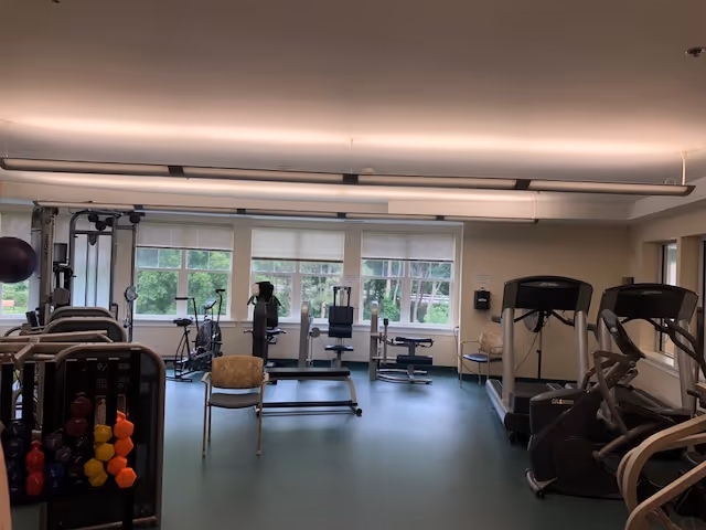 A fitness room with various exercise equipment including treadmills, stationary bikes, weight machines, and a rack of colorful dumbbells. The room has large windows letting in natural light and a chair placed near the center.