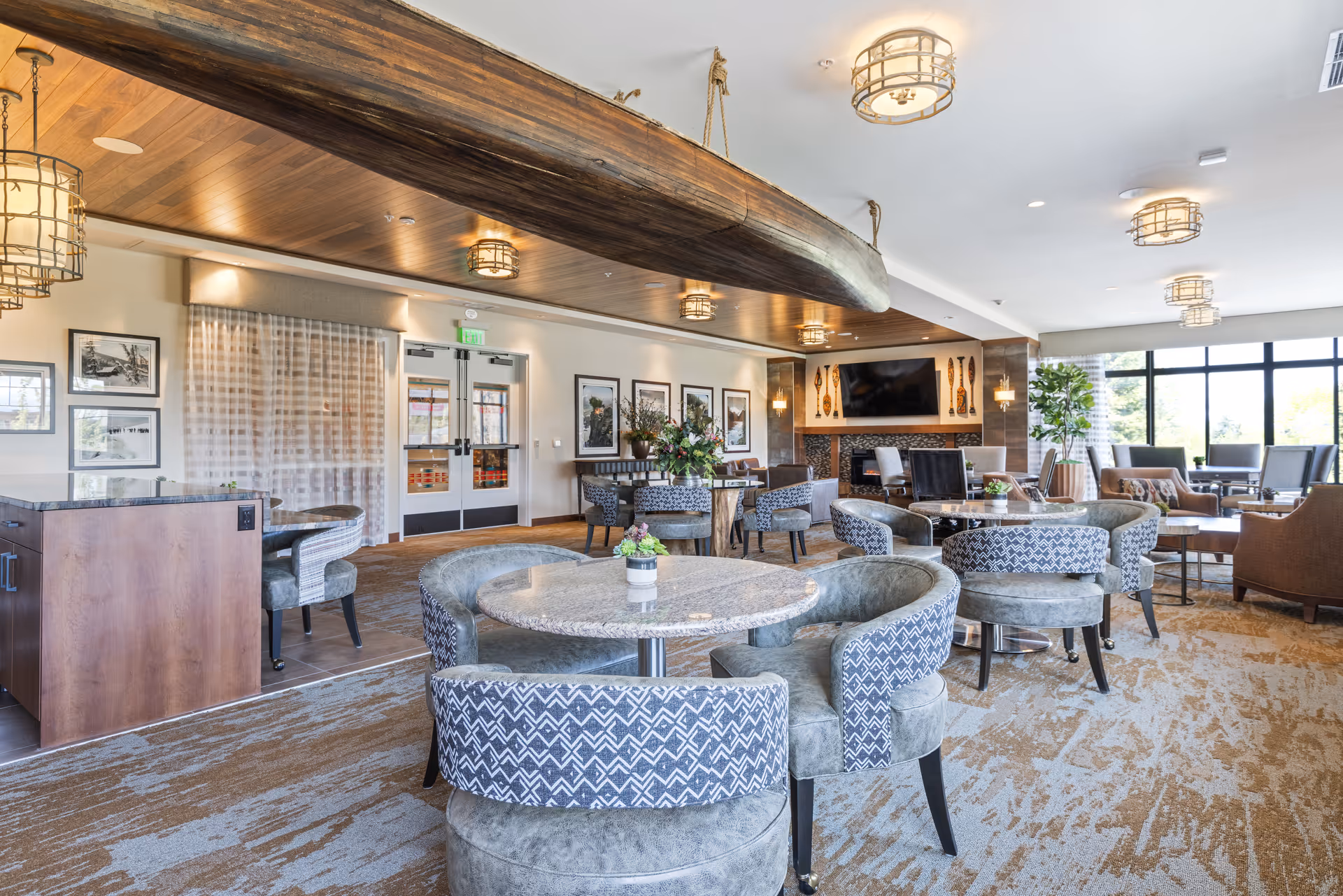 Bright, upscale common dining and lounge area with round stone-top tables, patterned upholstered chairs, a fireplace and TV, large windows, and a decorative wooden canoe suspended from the ceiling.