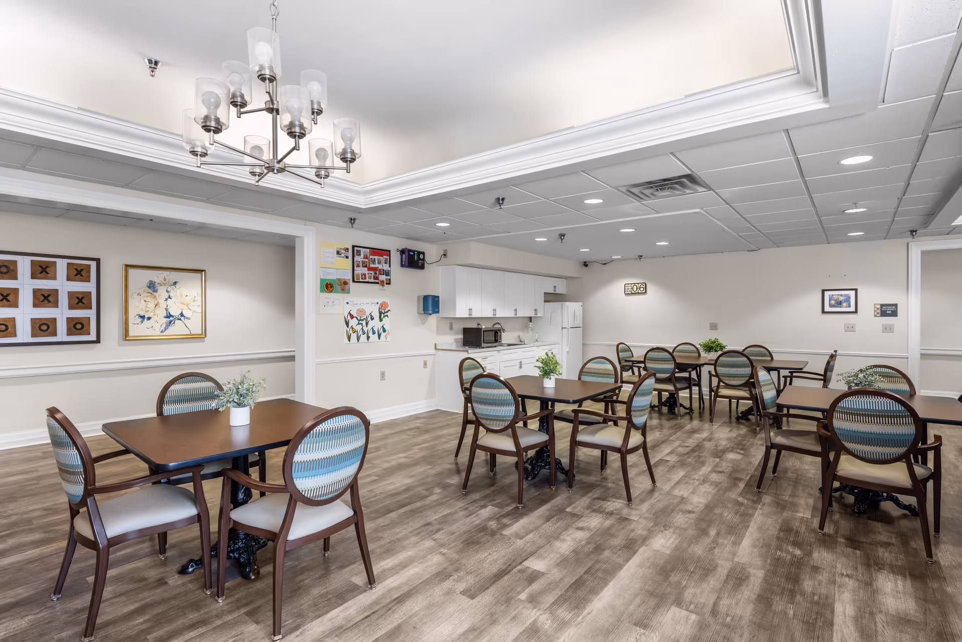 Communal dining room with several wooden tables and upholstered chairs, potted centerpieces, and a kitchenette against the far wall.