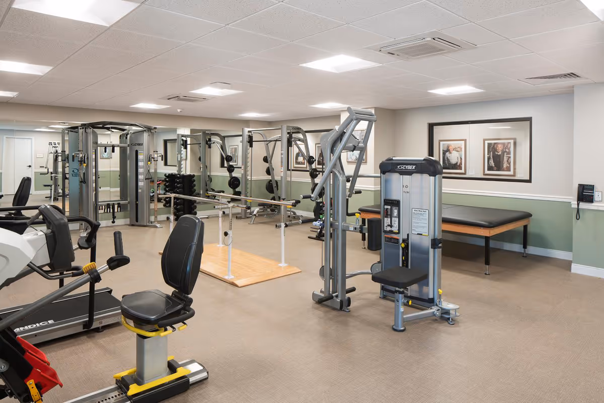 A well-lit senior living facility fitness room with various exercise equipment including a recumbent bike, weight machines, parallel bars for walking support, and a padded therapy table. The walls are painted light green and white, with framed pictures hanging on the wall and large mirrors reflecting the room.