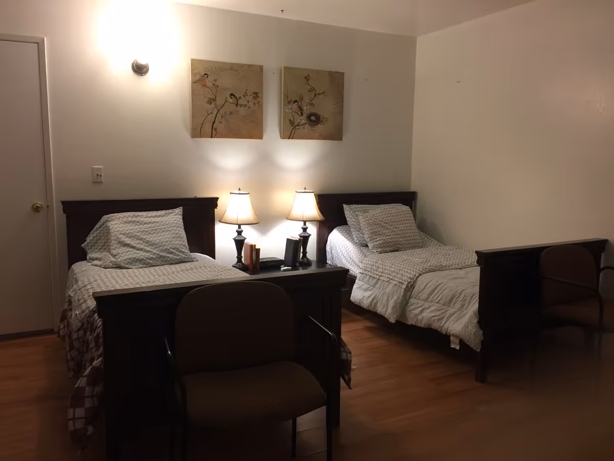 Two twin beds in a minimally decorated room with a shared nightstand, two lamps, chairs and wall art.