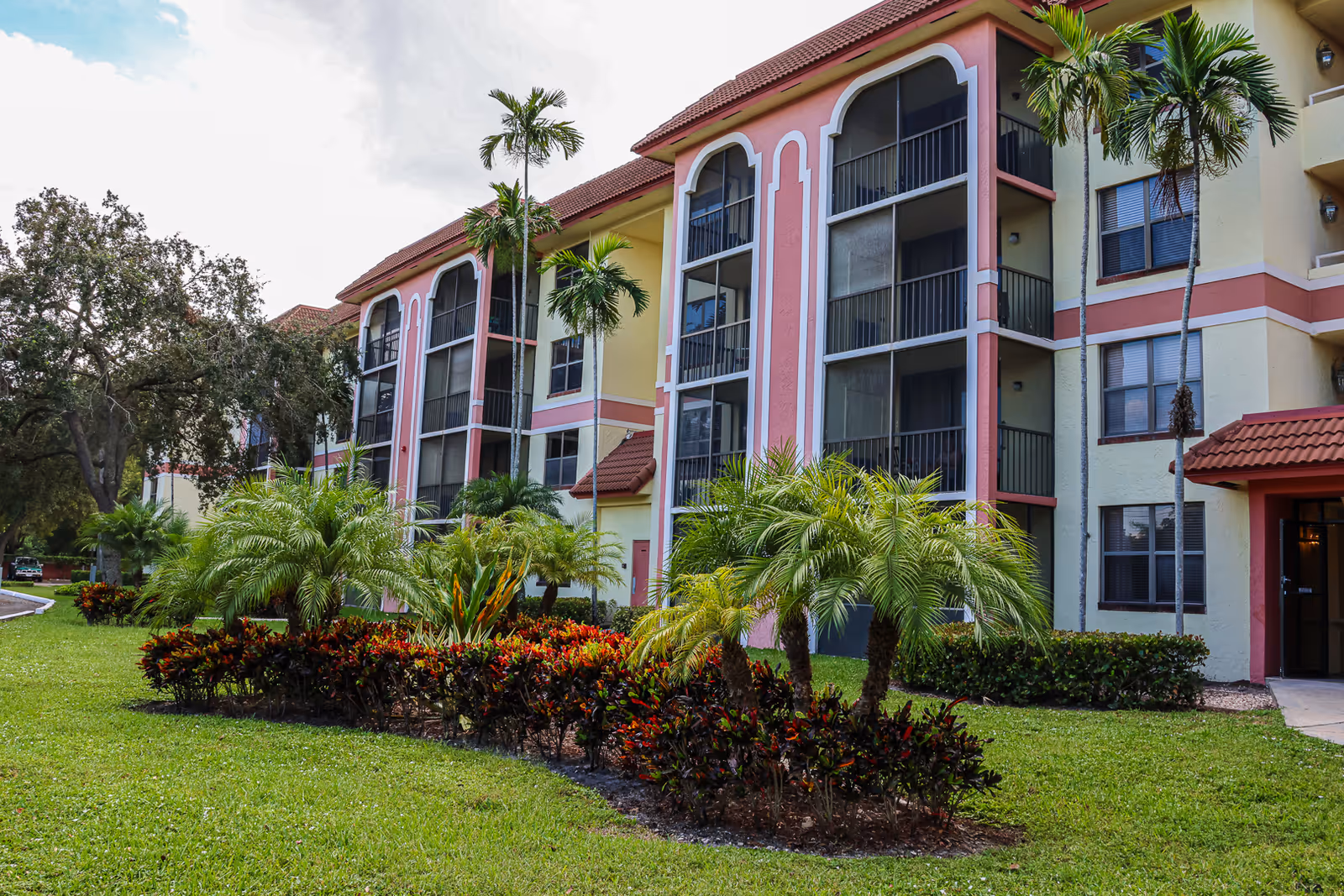 Exterior view of a multi-story residential building with screened balconies, palm trees, and landscaped greenery in front under a partly cloudy sky.