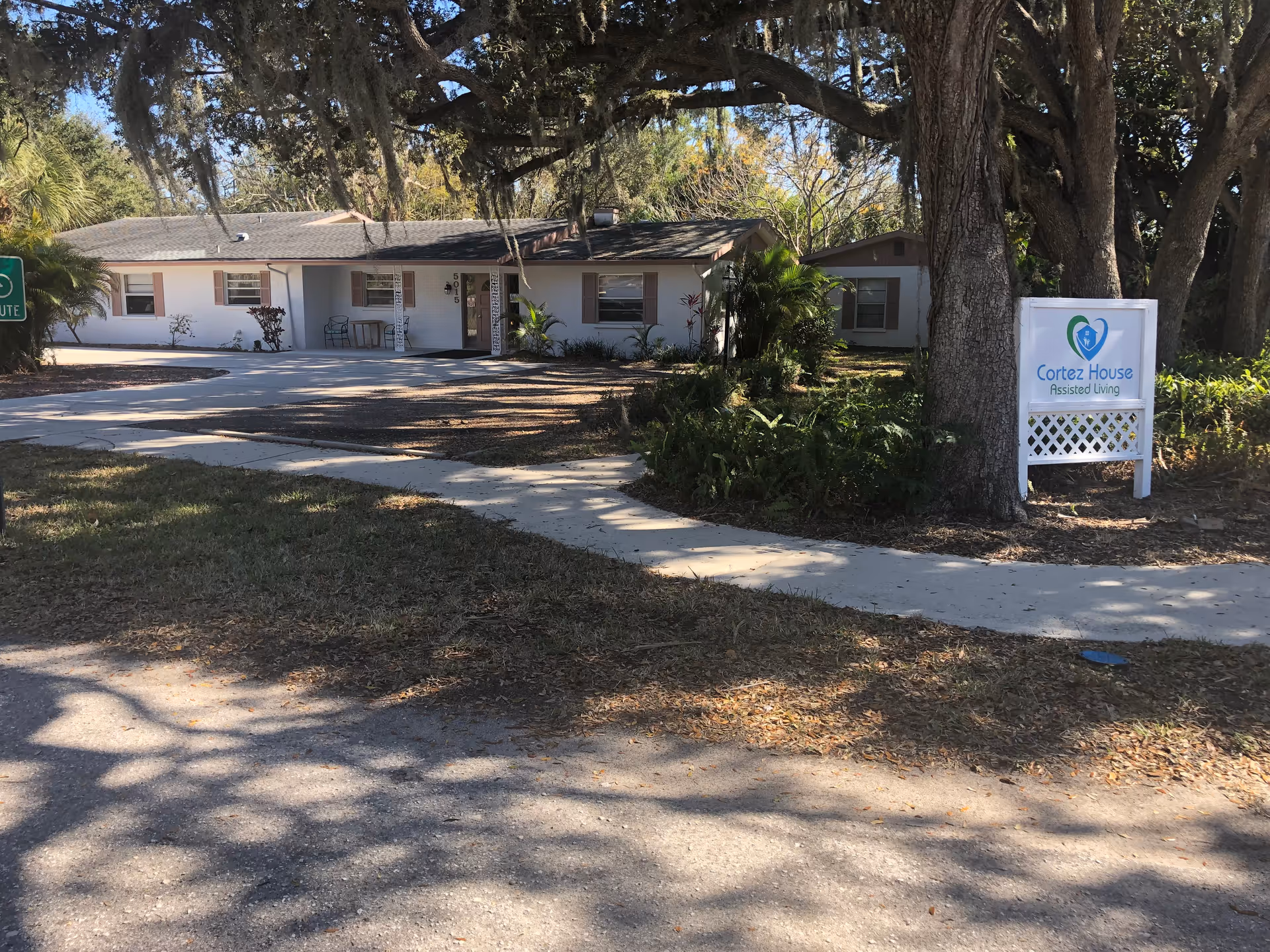 Exterior view of a single-story assisted living facility named Cortez House Inc, with a white building surrounded by trees and greenery. There is a sidewalk leading to the entrance and a sign in front displaying the facility's name and logo.