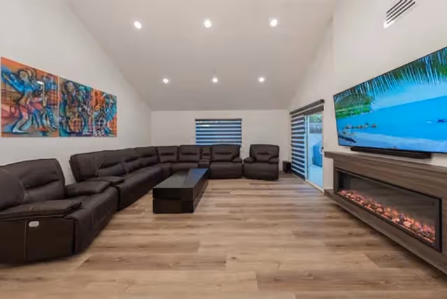 A modern, spacious living room with a long dark sectional sofa, a coffee table, a fireplace, and a wall-mounted TV in a senior living facility.