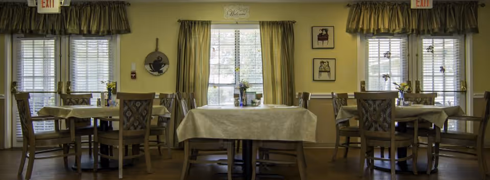 A dining room with several tables covered with beige tablecloths and wooden chairs arranged around them. The room has large windows with curtains allowing natural light to enter. There are decorative items on the walls and small flower vases on the tables. Two exit signs are visible above the doors on either side of the room.