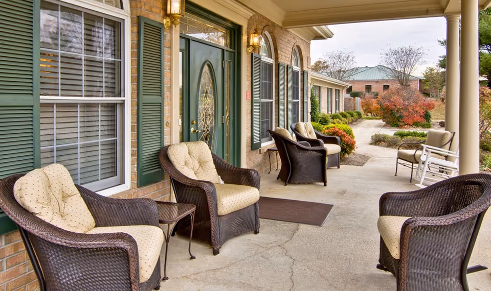 Outdoor covered patio area with cushioned wicker chairs and a small table in front of a brick building with green shutters and a green door, surrounded by landscaped bushes and trees with autumn foliage.