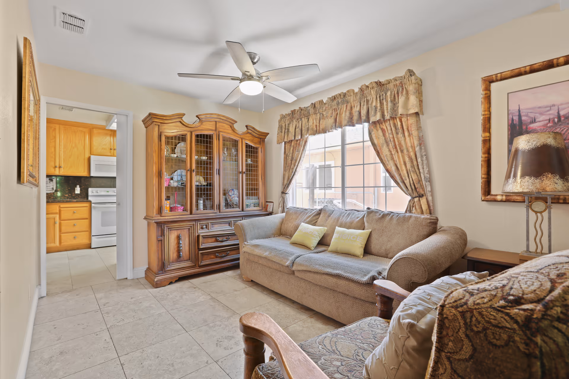 Cozy living room with a sofa, armchair, wooden china cabinet, ceiling fan, and a view into the kitchen through an open doorway.