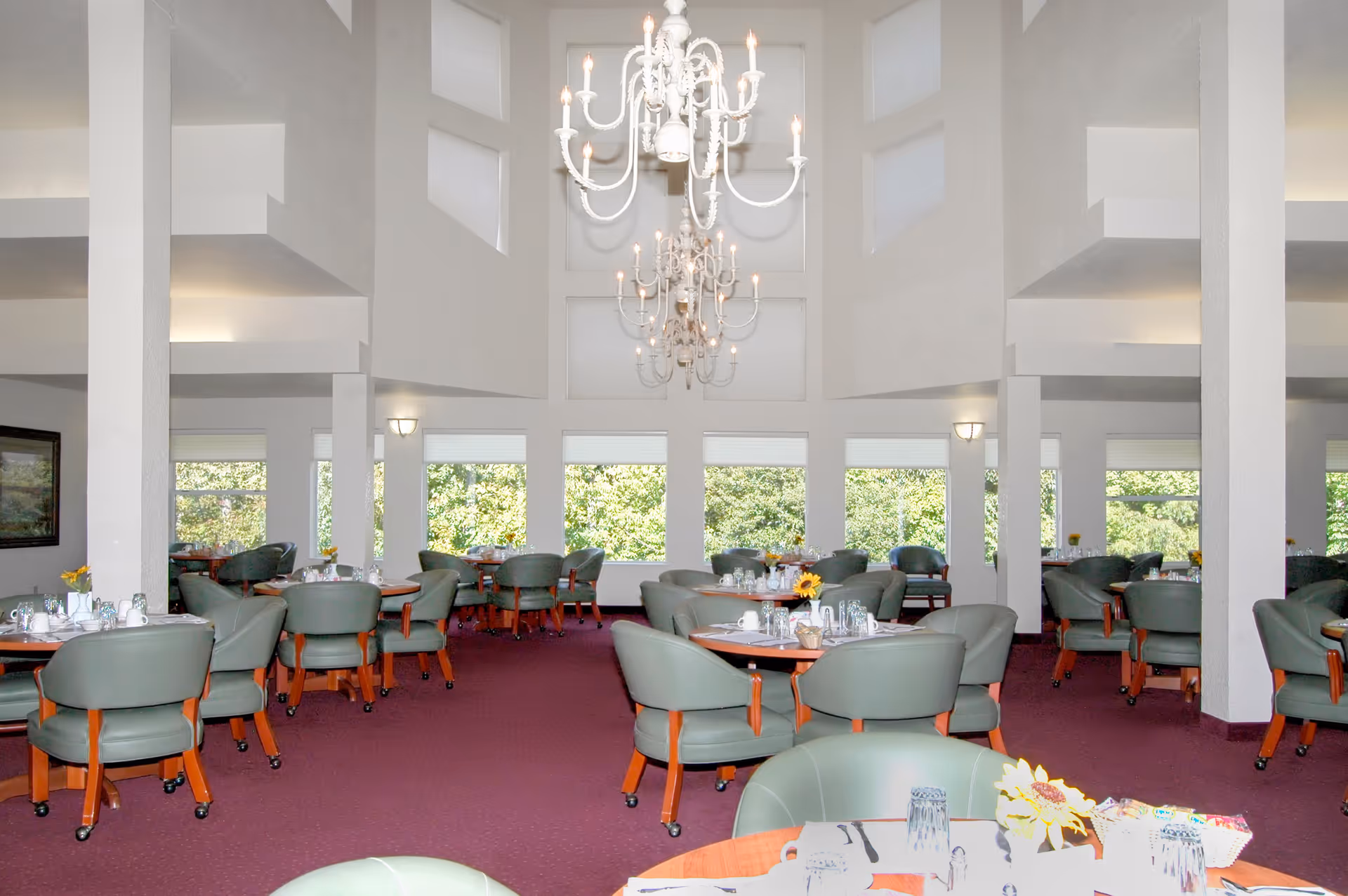 A spacious dining room with multiple round tables set with glasses, cups, and napkins. The room features green cushioned chairs with wooden legs, large windows letting in natural light, and two large white chandeliers hanging from a high ceiling. The carpet is a deep red color, and there are white walls with several pillars.