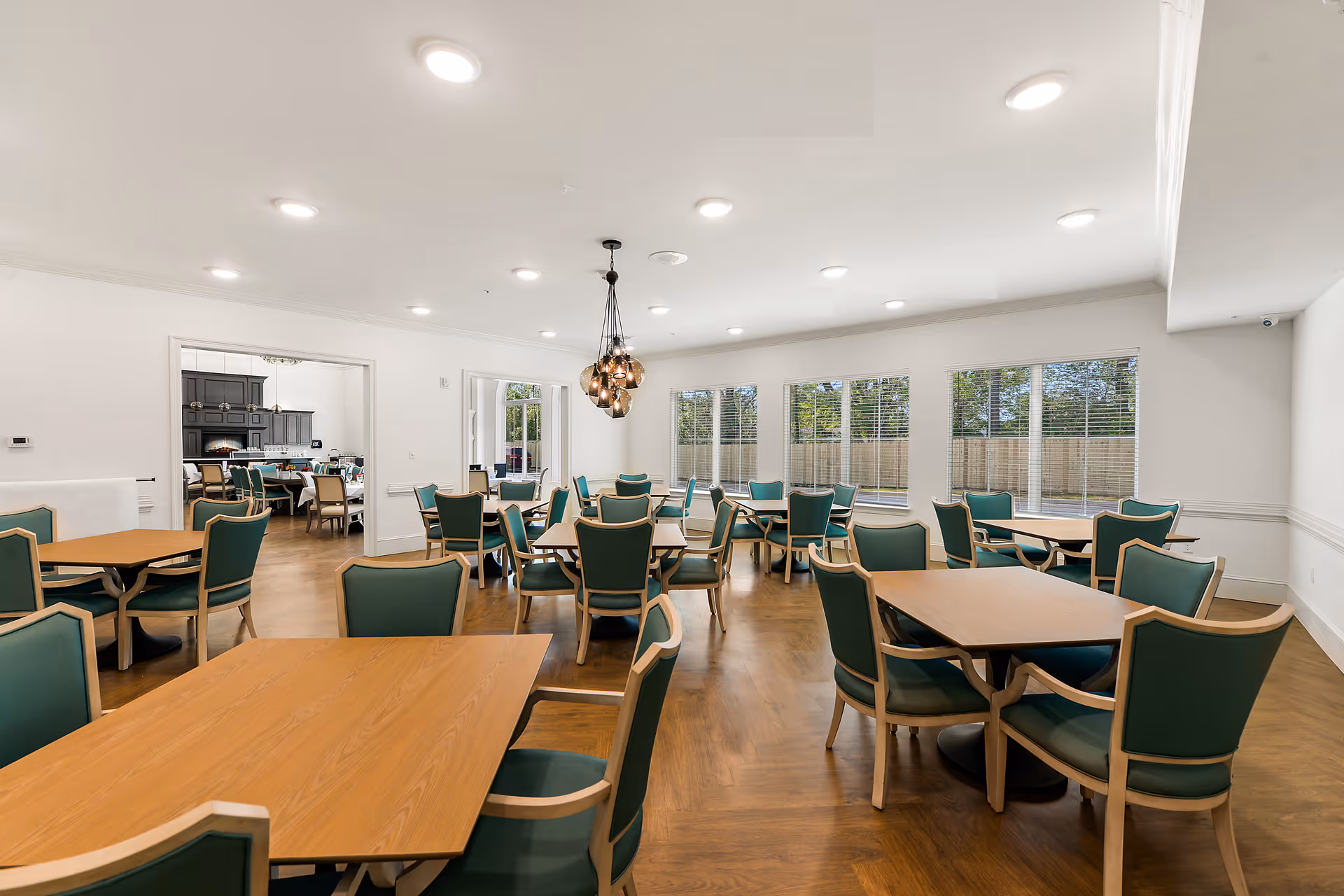 A spacious dining room with multiple wooden tables and green cushioned chairs arranged neatly. Large windows along one wall allow natural light to fill the room, and a modern chandelier hangs from the ceiling. The room has light-colored walls and a wooden floor, with an adjacent room visible through an open doorway.