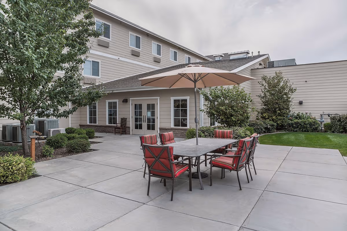 Patio courtyard with a large table, umbrella and red-cushioned chairs in front of a multi-story building.