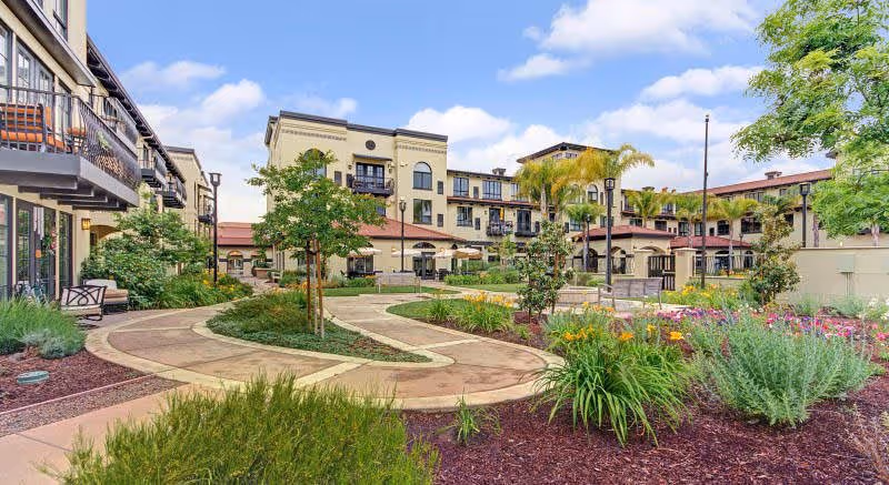 Outdoor garden area with winding paved pathways, landscaped greenery, colorful flowers, and several trees in front of a multi-story senior living facility building under a partly cloudy sky.