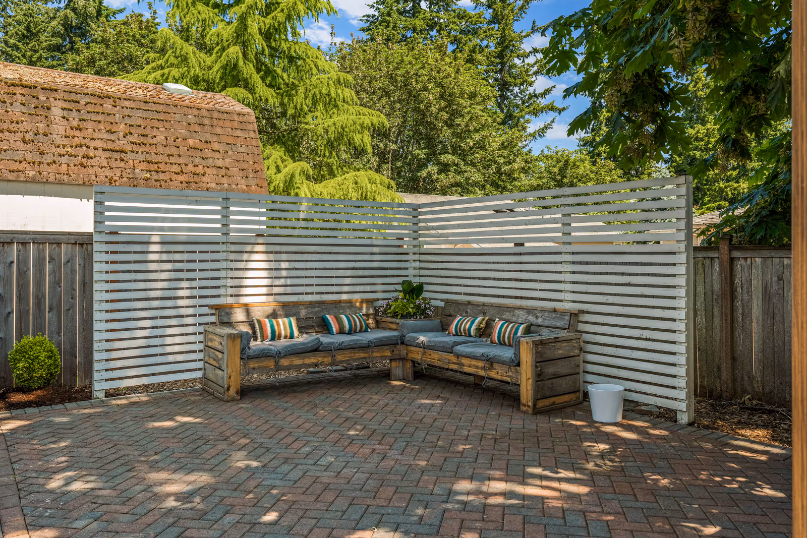 Outdoor patio area with a wooden L-shaped bench featuring gray cushions and striped pillows, surrounded by white slatted privacy fencing and tall green trees in the background.