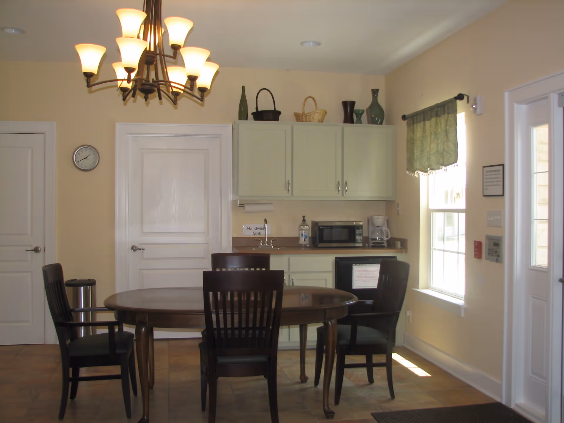 A small dining area with a round wooden table and four dark wooden chairs. Behind the table is a kitchenette with light green cabinets, a microwave, coffee maker, small sink, and a mini refrigerator. Above the cabinets are decorative vases and baskets. A chandelier with multiple lights hangs from the ceiling. There is a window with a green valance on the right side and two closed white doors on the left wall.