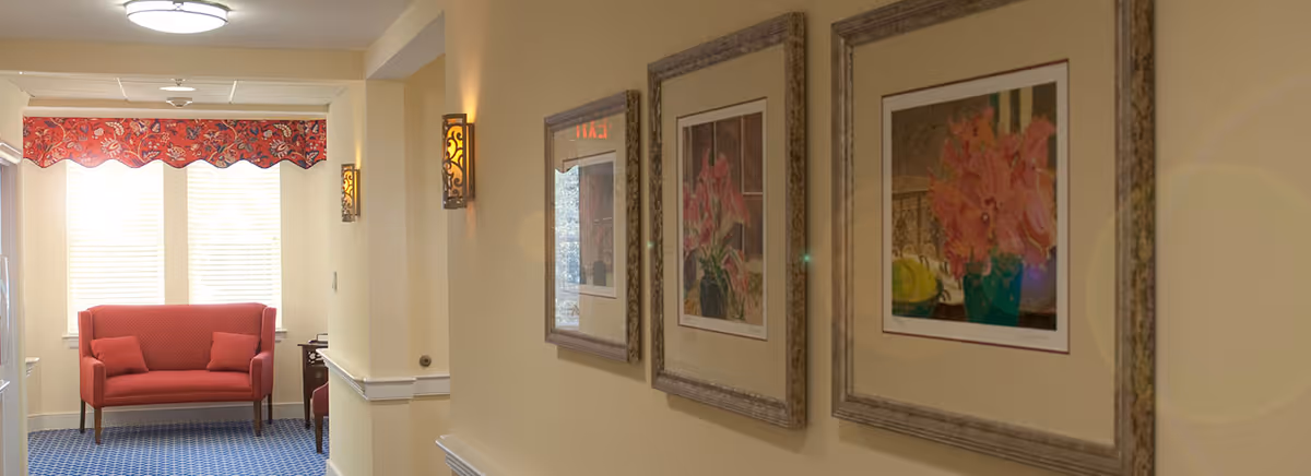 A hallway in a senior living facility with framed floral artwork on the right wall and a red loveseat with two cushions at the end near a window with a red valance. The floor is carpeted in blue with a pattern, and the walls are painted a light beige color with decorative wall sconces providing lighting.