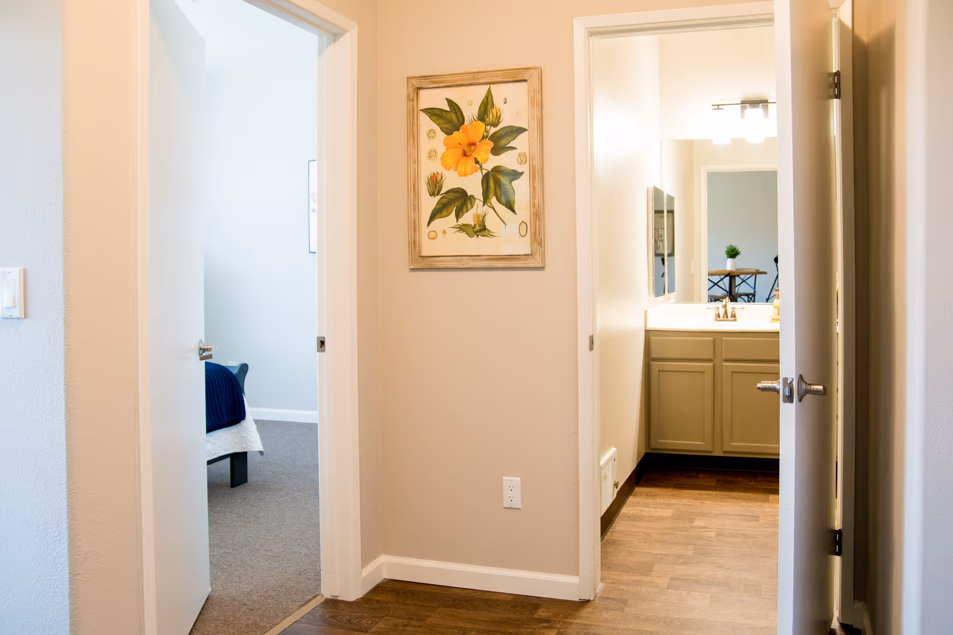 View of a hallway in a senior living facility with two open doors. One door leads to a bedroom with a bed partially visible, and the other door leads to a bathroom with a sink and mirror. A framed floral artwork is hung on the wall between the doors.
