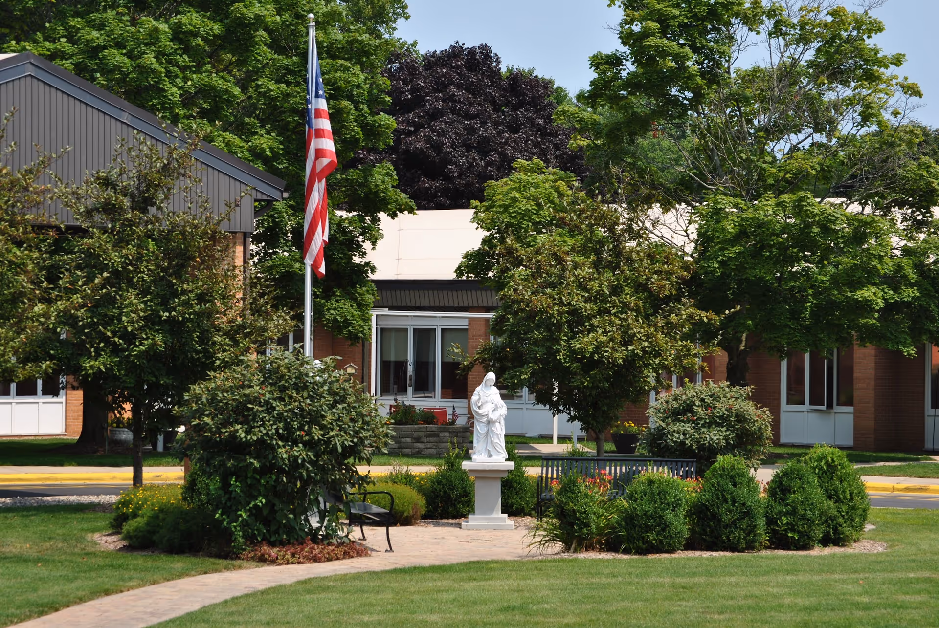 A landscaped outdoor area at St. Ann's Home featuring a white statue on a pedestal surrounded by bushes and trees. An American flag on a flagpole stands nearby, with a building visible in the background.