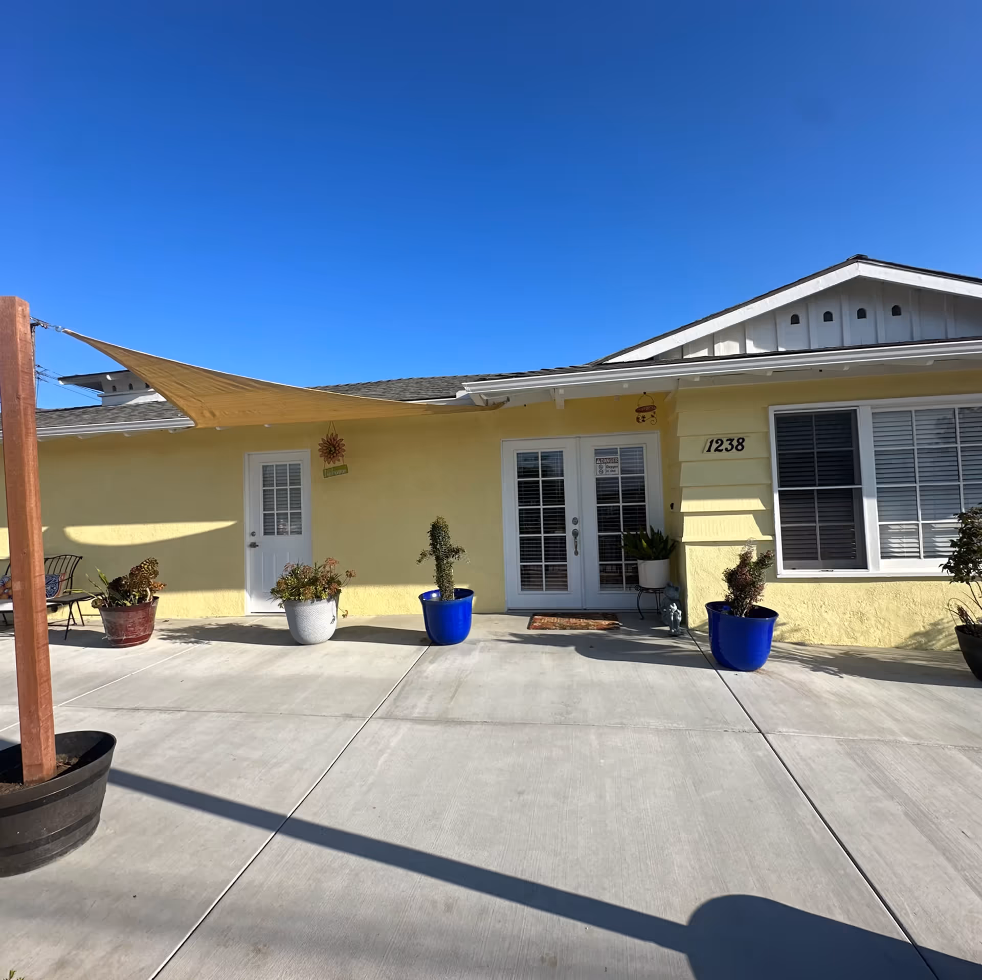Exterior view of a single-story yellow building with a concrete patio in front. The building has a white door and a set of white French doors with the number 1238 displayed on the wall. Several potted plants are placed along the patio, and a beige shade sail is stretched above part of the patio. The sky is clear and blue.
