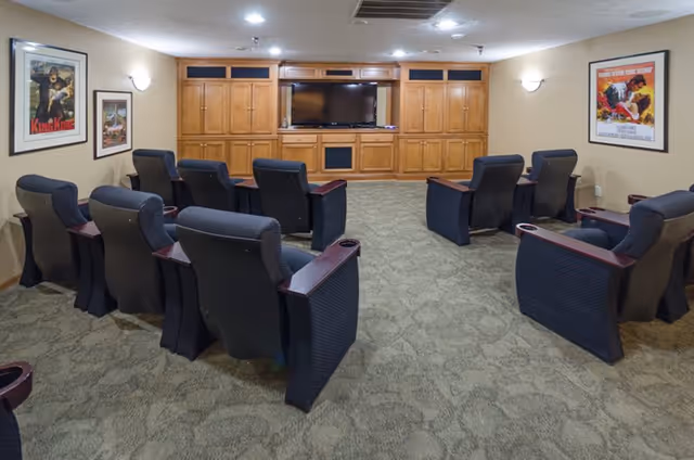 A small theater room with two rows of black cushioned recliner chairs facing a large flat-screen TV mounted on a wooden cabinet wall. The room has beige walls with framed movie posters and wall sconces providing light. The floor is carpeted with a patterned design.