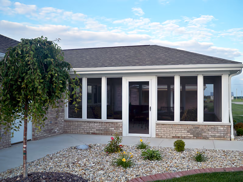 Exterior view of a single-story brick building with a screened-in porch. The porch has multiple large windows and a door. In front of the porch is a landscaped area with small plants, flowers, and decorative rocks. A small tree is visible on the left side of the image, and the sky is partly cloudy.