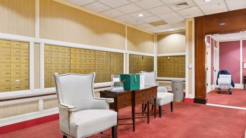 Interior view of a mailroom area with rows of gold mailboxes on the wall, two upholstered armchairs, a wooden table with a green box and some papers on it, red carpet flooring, and a hallway leading to another room with a cart and boxes.