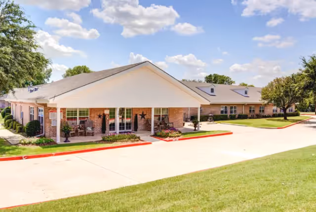 Exterior view of a single-story brick building with a covered entrance, decorated with star ornaments and potted plants. The building is surrounded by a well-maintained lawn, trees, and a paved driveway under a partly cloudy sky.