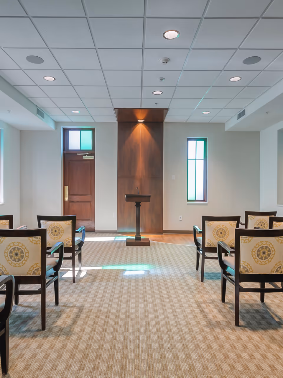 A small meeting or chapel room with a wooden podium centered against a wood-paneled wall. The room has beige patterned carpet, several chairs with decorative backs arranged in rows facing the podium, and two narrow windows on either side of the podium letting in natural light.