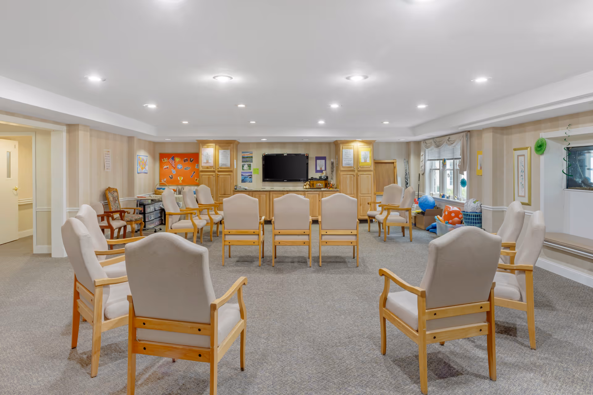 A spacious common room with beige carpet and walls, featuring multiple beige cushioned wooden chairs arranged in a circle. The room has a flat-screen TV mounted on a wooden cabinet against the far wall, bulletin boards, and windows with valances letting in natural light. Various decorations and storage bins are visible near the windows.
