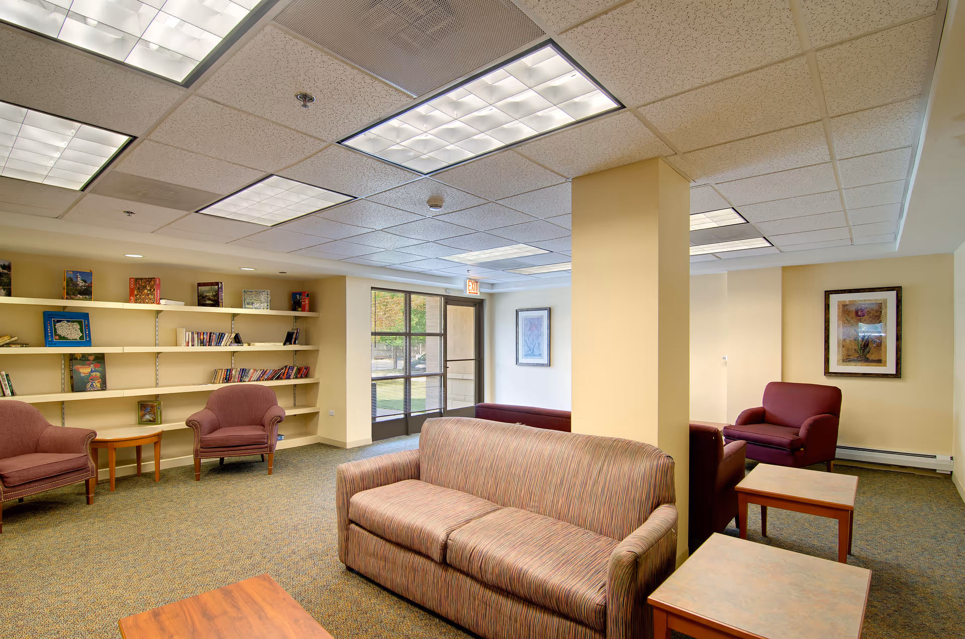 A well-lit senior living facility common area with multiple upholstered chairs and sofas arranged around wooden tables. There are built-in shelves on one wall holding books and decorative items, and framed artwork on the walls. Large windows and a glass door provide natural light and a view of the outdoor greenery.