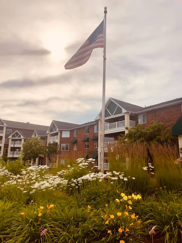 A senior living facility building with red brick exterior and white balconies, surrounded by a garden with green plants and yellow and white flowers. An American flag is flying on a flagpole in front of the building under a cloudy sky.