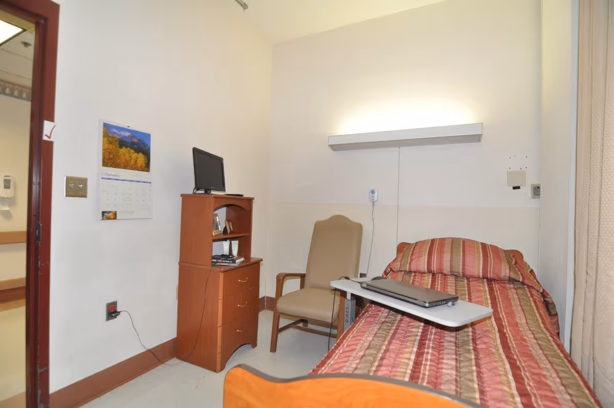 A small room with a single bed covered in a striped red and beige blanket. Next to the bed is a beige armchair and a small wooden dresser with a TV on top. A calendar is hanging on the wall, and a light fixture is mounted above the bed. The room has plain white walls and a door leading to a hallway.