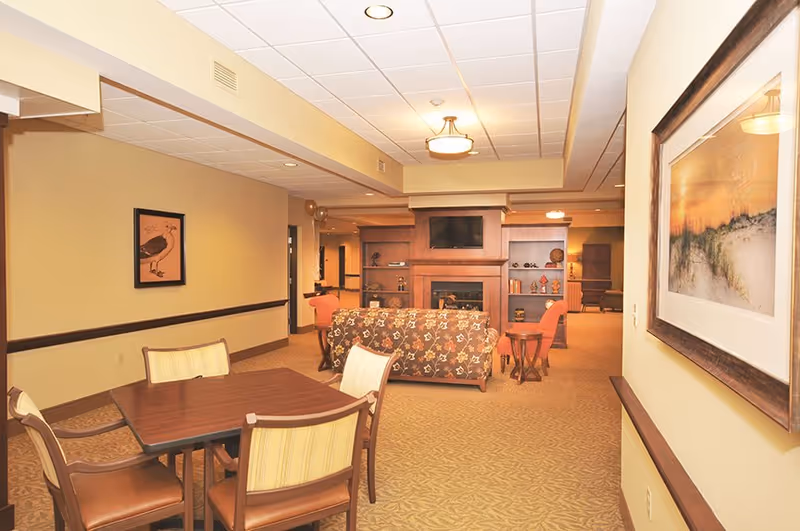 A warm furnished common area with a dining table in the foreground and a seating area with a fireplace and TV in the background.