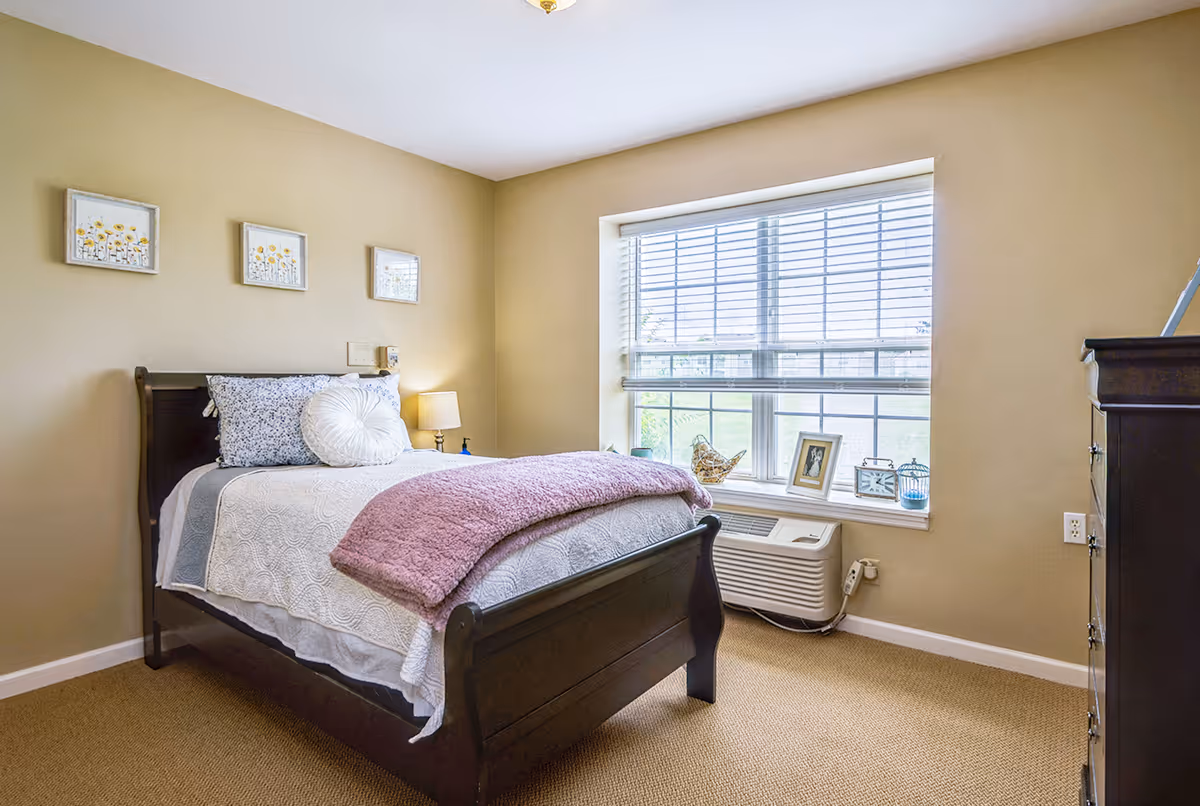 A cozy bedroom with a single dark wooden bed frame, white and blue bedding, and a purple throw blanket. There is a window with blinds letting in natural light, a small lamp on a nightstand, and three framed pictures of flowers on the beige wall. A dark wooden dresser is partially visible on the right side.