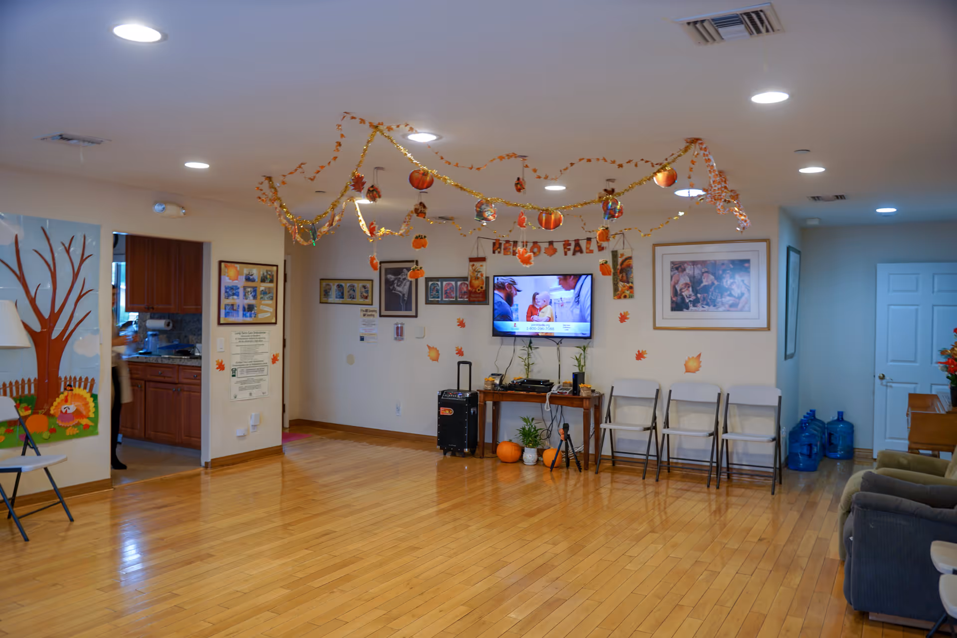 A spacious room with wooden flooring decorated with fall-themed garlands and hanging ornaments. A flat-screen TV is mounted on the wall displaying a program, with a small table below holding plants and pumpkins. Folding chairs are lined up against the wall, and there are framed pictures and posters on the walls. To the left, there is an open doorway leading to a kitchen area with wooden cabinets. The room is well-lit with ceiling lights.