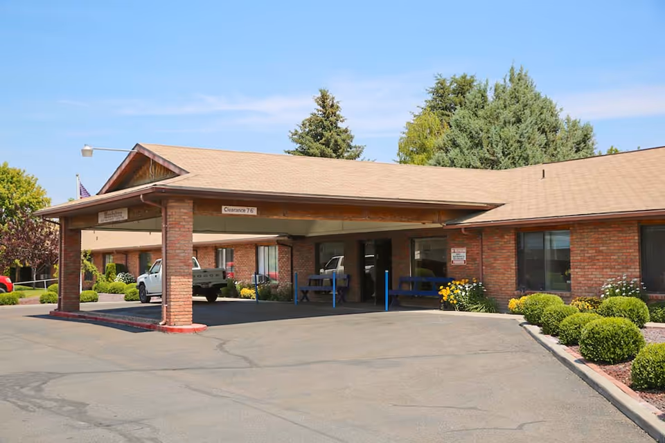 Exterior view of Woodstone Assisted Living facility showing a covered entrance with brick pillars, a driveway, parked vehicles, and landscaped bushes and trees under a clear blue sky.